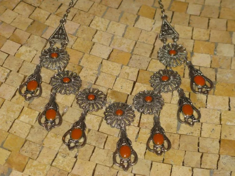 Ce collier en métal argenté, agrémentée de breloques en forme de fleurs et de pendentifs en pierre orange vif.